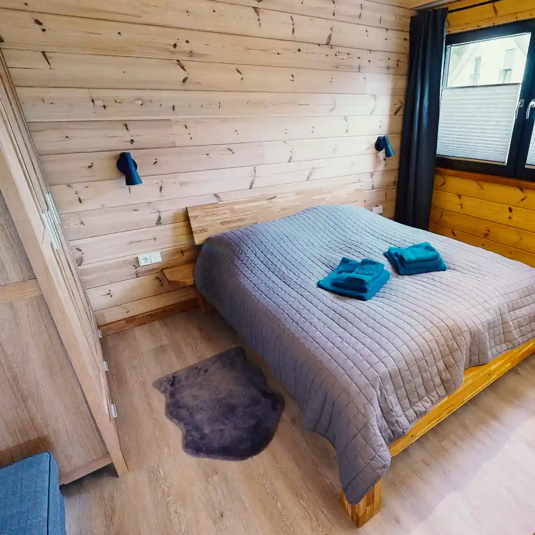Ein Standbild aus einer virtuellen Tour von raum media, entstanden für das Ferienhaus Eifelspur. Zu sehen ist das Schlafzimmer im Ferienhaus Eifelspur in Netterhseim, ausgestattet mit einem Doppelbett mit grauer Bettdecke und blauen Handtüchern, Holzverkleidung an den Wänden, einem Fenster, das Tageslicht hereinlässt, und einem gemütlichen Teppich auf dem Boden.