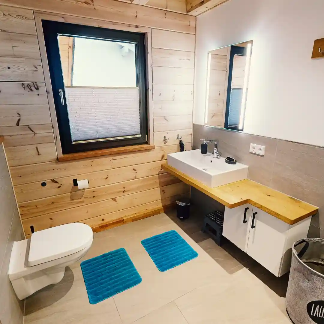 Ein Standbild aus einer virtuellen Tour von raum media, entstanden für das Ferienhaus Eifelspur. Zu sehen ist ein Badezimmer im Ferienhaus Eifelspur in Netterhseim mit modernen Elementen, einem Waschbecken auf einer Holzablage, einem großen Spiegel, einer Toilette und blauen Badematten auf dem Boden, umgeben von Holzvertäfelung und einem Fenster, das für Tageslicht sorgt.