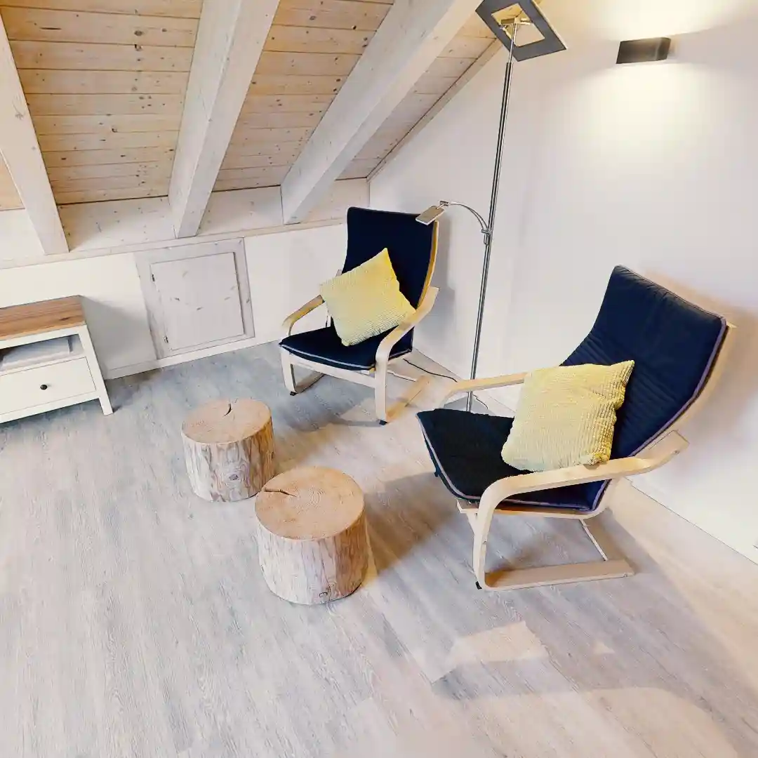 Ein Standbild aus einer virtuellen Tour von raum media, entstanden für das Ferienhaus Eifelspur. Zu sehen ist eine Gemütliche Sitzecke im Ferienhaus Eifelspur in Netterhseim, bestehend aus zwei modernen Stühlen mit blauen Polstern und gelben Kissen, begleitet von zwei rustikalen Holzscheiben als Tische, unter einem schrägen Holzdecke und einer Stehlampe.