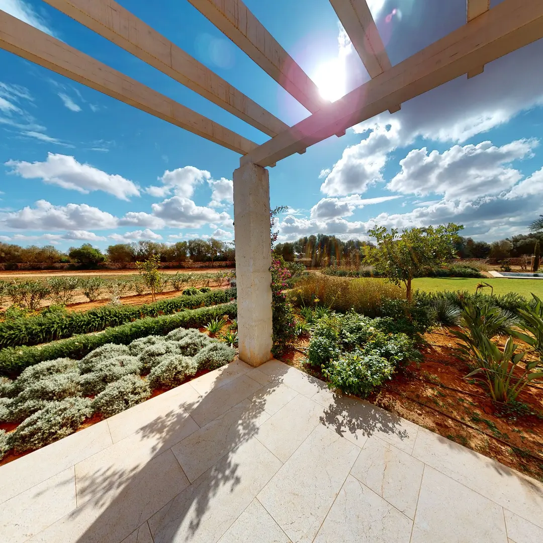 Ein Standbild aus einer virtuellen Tour von raum media, entstanden für die Penta Gruppe. Zu sehen ist die Landschaft von einer überdachten Terrasse in einer Finca auf Mallorca, umgeben von üppigem Grün, Sträuchern und Bäumen, unter einem strahlend blauen Himmel mit Wolken und der Sonne, die durch eine Pergola scheint.