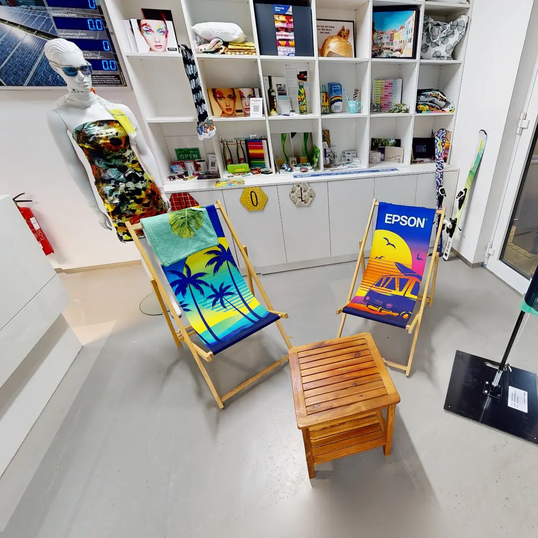 Ein Standbild aus einer virtuellen Tour von raum media, entstanden für Epson. Zu sehen sind Zwei liegestühle mit bunten Aufdrucken in einem modernen Raum des Epson Industrial Solutions Centers, umgeben von Regalen mit verschiedenen Produkten und Dekorationen. Ein Liegestuhl zeigt eine tropische Landschaft mit Palmen und Sonnenuntergang, während der andere den Schriftzug "Epson" trägt. In der Mitte befindet sich ein kleiner Holztisch. Im Hintergrund ist eine Schaufensterpuppe in einem modischen Kleid zu sehen.