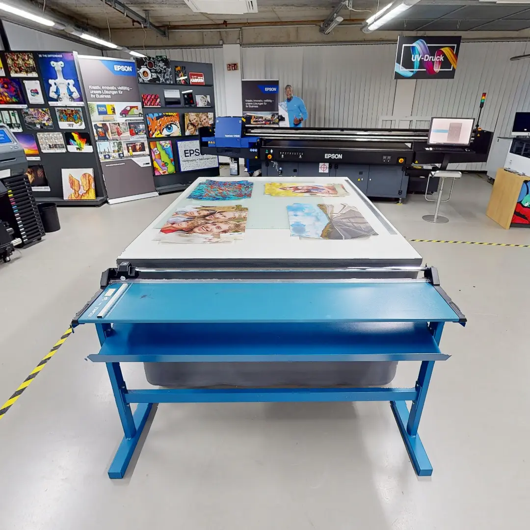 Ein Standbild aus einer virtuellen Tour von raum media, entstanden für Epson. Zu sehen ist eine moderne Druckumgebung im Epson Industrial Solutions Center, die einen großen Flachbettdrucker zeigt. Auf dem Drucker liegt ein bedrucktes Material mit verschiedenen farbigen Grafiken, darunter Porträts und künstlerische Designs. Im Hintergrund sind weitere Epson-Drucker und eine Informationswand mit verschiedenen Druckmustern und -techniken zu sehen. Ein Mitarbeiter in blauer Kleidung steht in der Nähe und beobachtet den Druckprozess. Die Umgebung ist hell beleuchtet und gut organisiert, mit einem sauberen, industriellen Eindruck.