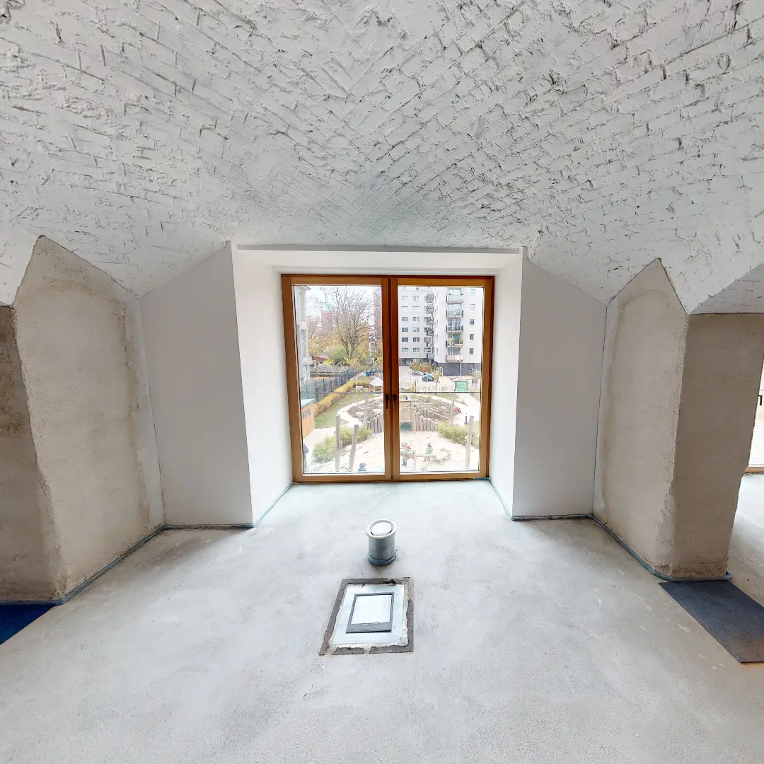 Ein Standbild aus einer virtuellen Tour von raum media, entstanden für die PANDION GmbH. Zu sehen ist die Innenansicht eines zukünftigen Büros in Berlin mit einem großen Fenster, das einen Blick auf einen Garten und umliegende Gebäude bietet. Der Raum hat eine helle, schlichte Gestaltung mit einem gewölbten, weißen Ziegeldach und einem unvollendeten Boden. Ein zylindrischer Behälter steht in der Mitte des Raumes, während die Wände teilweise verputzt sind.