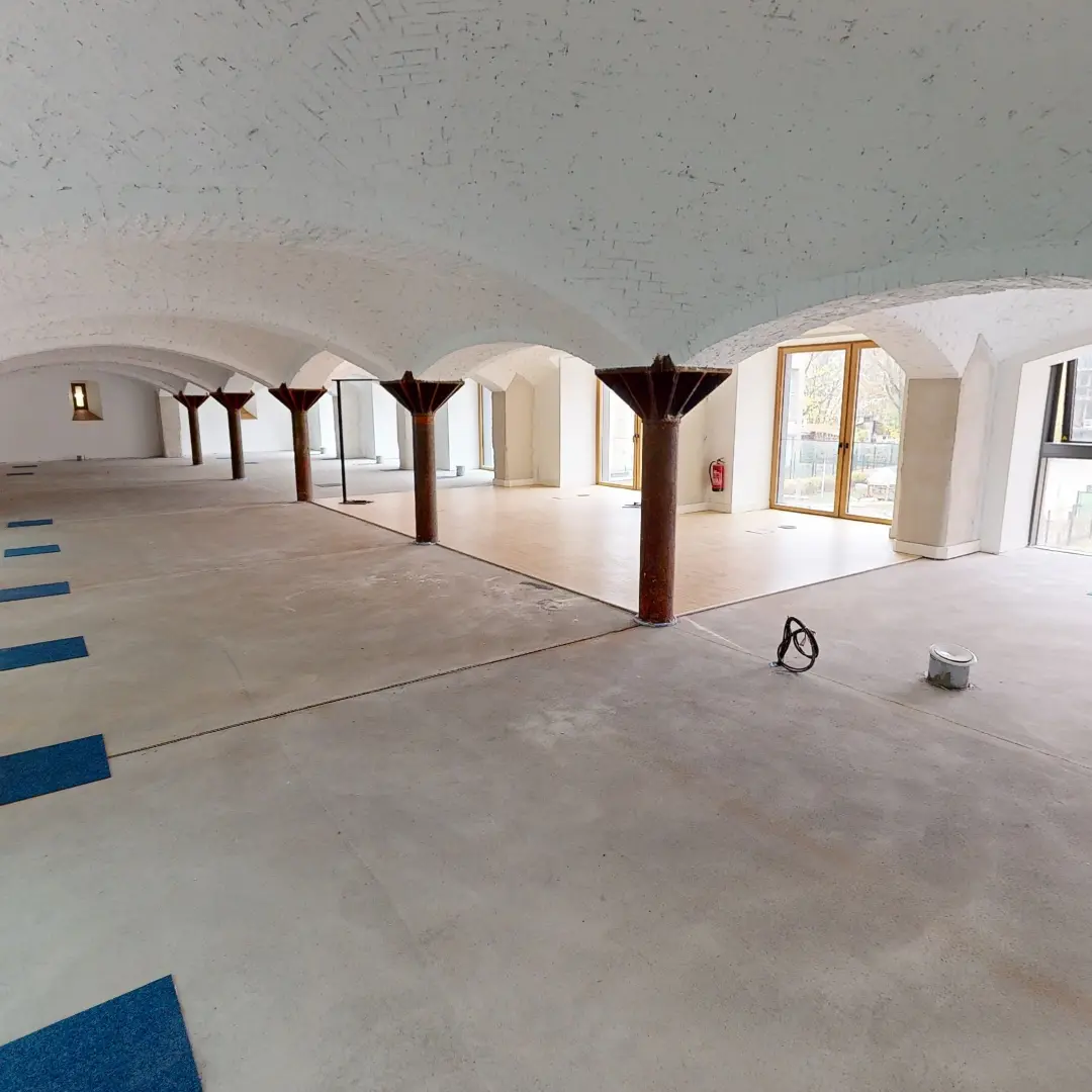Ein Standbild aus einer virtuellen Tour von raum media, entstanden für die PANDION GmbH. Zu sehen ist ein großer, offener Büroraum in Berlin mit hohen, gewölbten Decken und architektonischen Säulen. Der Raum verfügt über große Fenster, die viel natürliches Licht hereinlassen, und ist teilweise mit blauen Teppichen ausgelegt. Der Boden ist teilweise uneben und zeigt verschiedene Materialien.