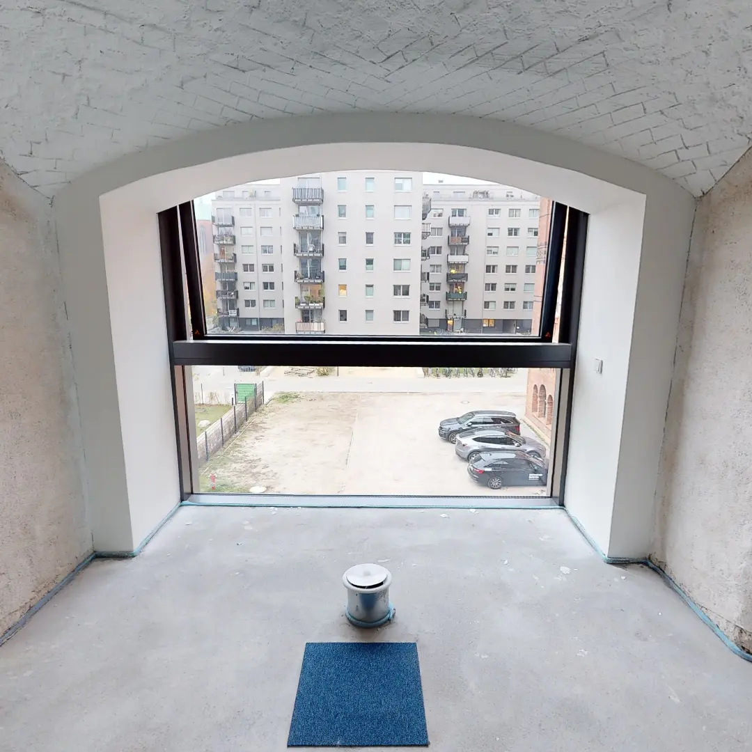 Ein Standbild aus einer virtuellen Tour von raum media, entstanden für die PANDION GmbH. Zu sehen ist die Innenansicht eines zukünftigen Büros in Berlin mit einem großen Fenster, das einen Blick auf einen Garten und umliegende Gebäude bietet. Der Raum hat eine helle, schlichte Gestaltung mit einem gewölbten, weißen Ziegeldach und einem unvollendeten Boden. Ein zylindrischer Behälter steht in der Mitte des Raumes, während die Wände teilweise verputzt sind.