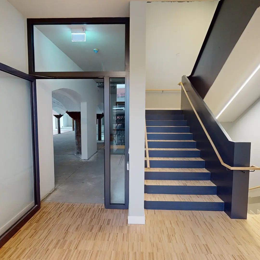 Ein Standbild aus einer virtuellen Tour von raum media, entstanden für die PANDION GmbH. Zu sehen ist der Eingangsbereich eines modernen Büros in Berlin, mit einem klaren Blick auf eine Treppe aus Holz und Metall. Der Boden ist mit einem hellen Holzbelag versehen, während die Wände hell gestrichen sind. Durch große Fenster auf der linken Seite strömt Tageslicht in den Raum, und im Hintergrund sind weitere Büros sichtbar.