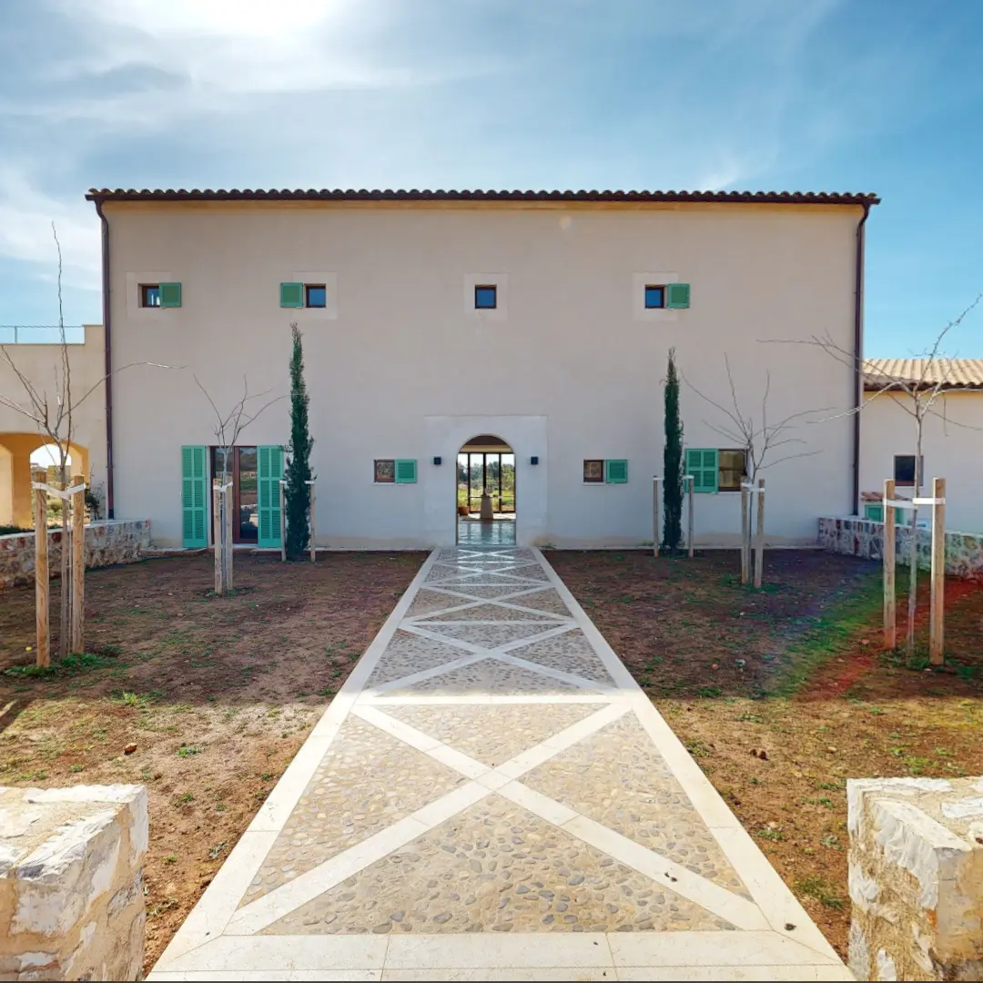 Ein Standbild aus einer virtuellen Tour von raum media, entstanden für die Penta Gruppe. Zu sehen ist der Eingang zu einer modernen Finca in Mallorca, mit einem gewölbten Tor aus Holz, das auf einen hellen Innenraum mit einem runden Teppich, einem Tisch und Stühlen blickt. Links steht ein großer Terrakotta-Blumentopf mit einer Pflanze, während die Wände in sanften Erdtönen gehalten sind.