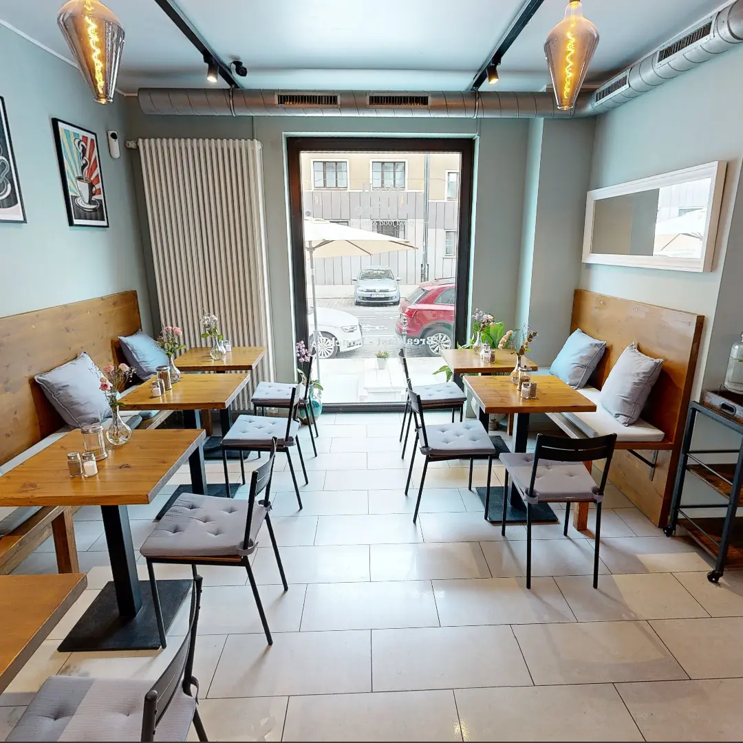 Ein Standbild aus einer virtuellen Tour von raum media, entstanden für die PANDION GmbH. Zu sehen ist ein großer, offener Büroraum mit gewölbten Decken und einer Reihe von Säulen. Die Wände sind in hellem Weiß gehalten und es gibt große Fenster, die für natürliches Licht sorgen. Die Säulen sind aus Metall und teilweise mit gelben und schwarzen Streifen markiert. Der Boden ist grau und teilweise mit blauen Fliesen akzentuiert. Der Raum vermittelt eine moderne und großzügige Atmosphäre, ideal für die Nutzung als Bürofläche.