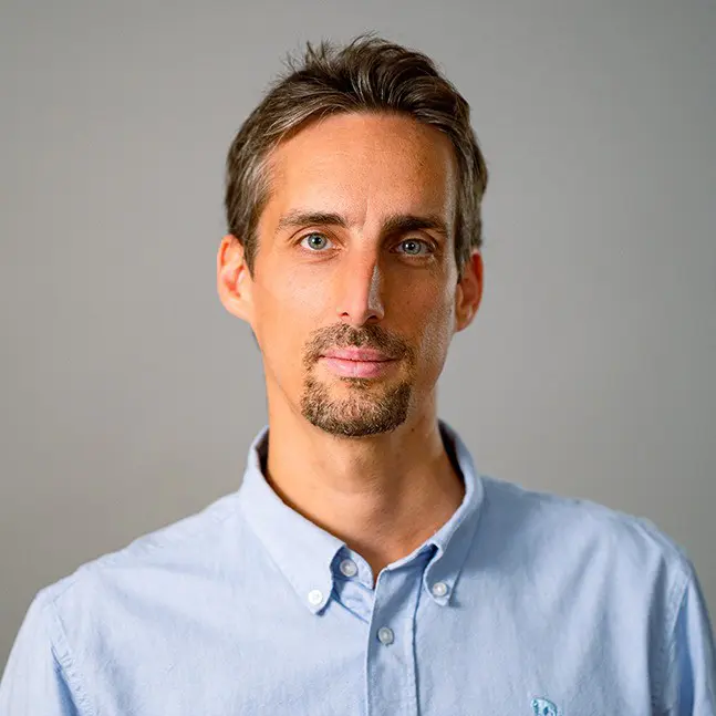 Porträt von Julian Mende, Leiter Produktion bei raum media, mit kurzem, graubraunem Haar und einem gepflegten Bart, der in einem hellblauen Hemd vor einem neutralen Hintergrund steht.