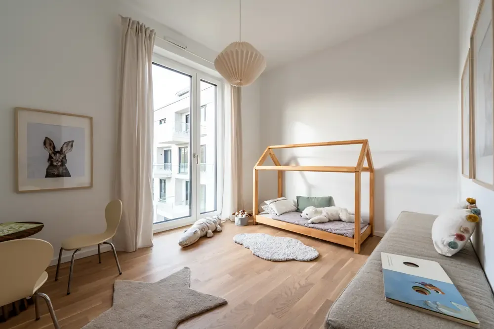 Immobilienfotos eines Kinderzimmers in einer Wohnung am Albertussee Düsseldorf, mit einem Holzbett im Hausdesign, einem gemütlichen Teppich, einem kleinen Tisch mit Stühlen und großen Fenstern, die viel Tageslicht einlassen.