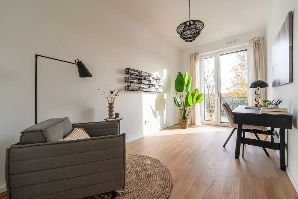 Wohnung am Albertussee Düsseldorf, modernes Wohnzimmer mit grauem Sofa, Stehlampe, Schreibtisch und großen Fenstern, die viel Tageslicht hereinlassen, dekoriert mit einer Pflanze und einem Wandregal.
