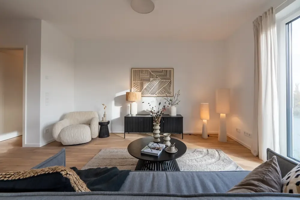 Wohnzimmer in einer Wohnung am Albertussee Düsseldorf, eingerichtet mit einem runden Couchtisch, einer gemütlichen Couch, einem modernen Teppich und dekorativen Lampen. Im Hintergrund steht ein Sideboard mit einer Wanddekoration.