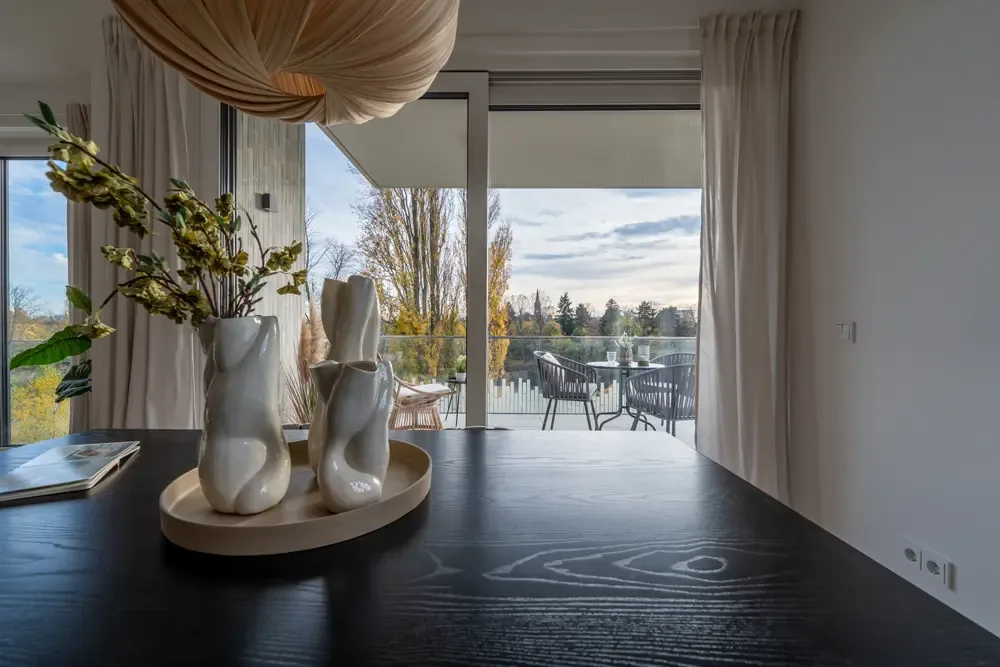 Innenansicht einer modernen Wohnung am Albertussee in Düsseldorf, mit einem Holztisch, dekorativen Vasen und Blick auf den Balkon mit Gartenmöbeln und Blick auf die Landschaft.