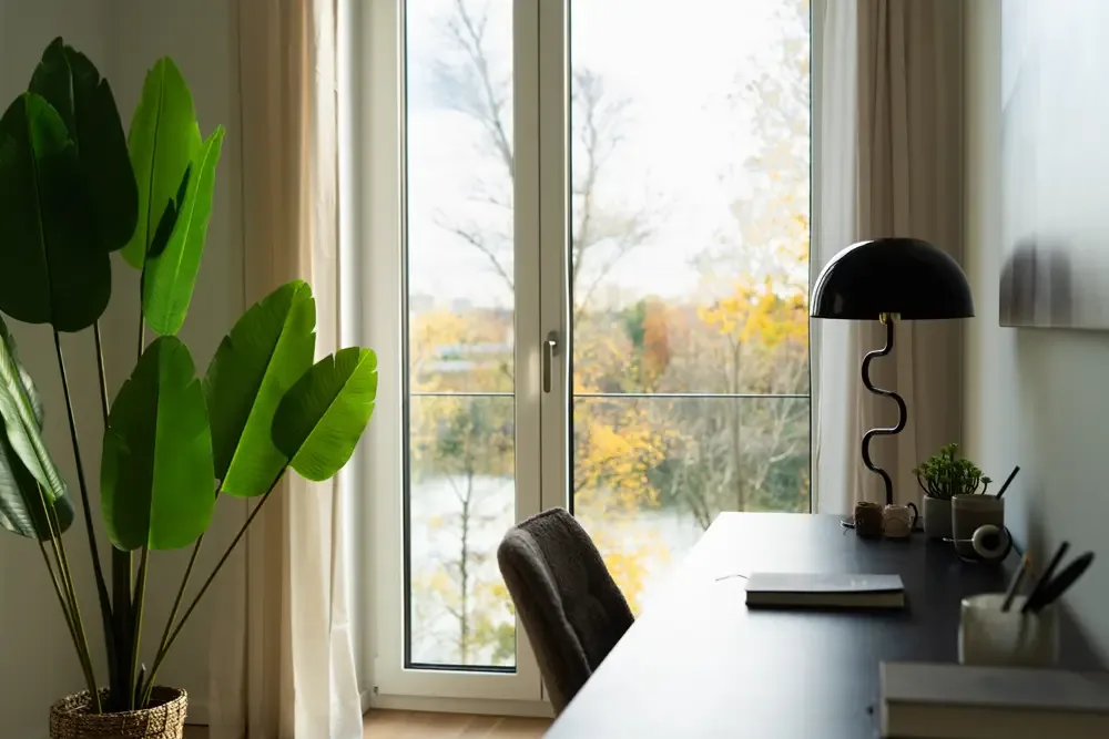 Ein modernes Arbeitszimmer mit einem Schreibtisch, einer schwarzen Lampe und einem Stuhl, im Hintergrund große Fenster mit Blick auf den Albertussee in Düsseldorf und einer Pflanze im Vordergrund.