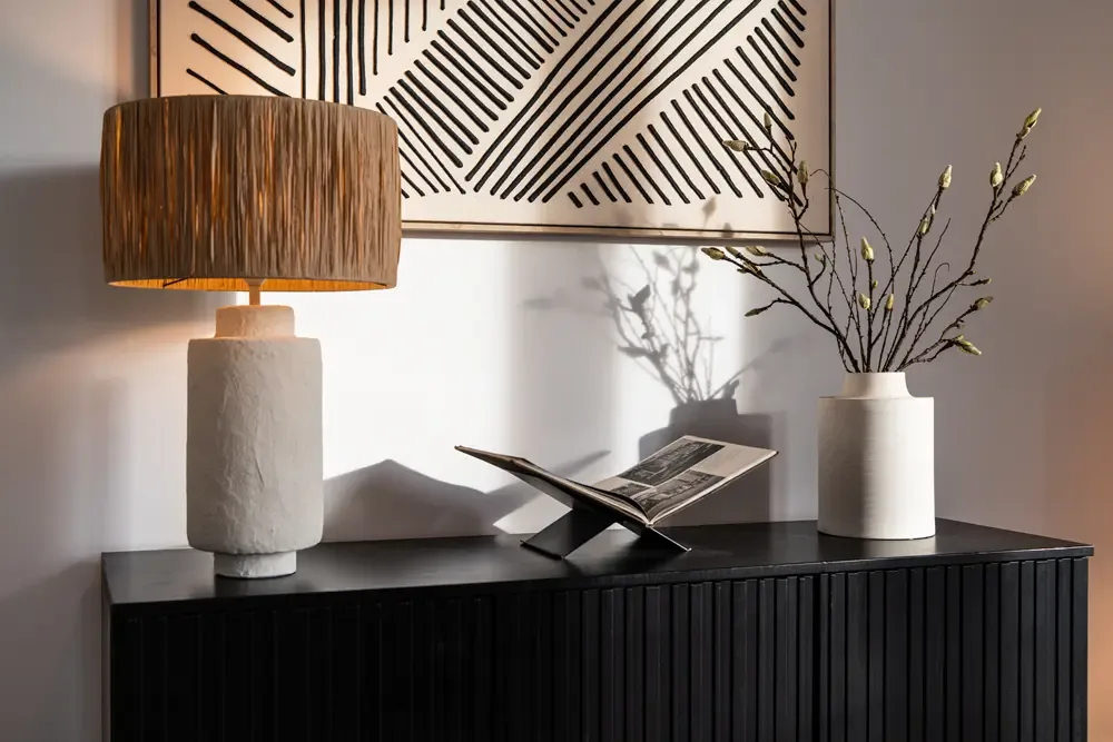 Ein moderner Tisch mit einer weißen Lampe und einem Lampenschirm aus geflochtenem Material, einem Buchständer mit einem geöffneten Buch, einer weißen Vase mit Zweigen und einer geometrischen Wanddekoration im Hintergrund, ideal für die Einrichtung von PANDION GmbH am Albertussee in Düsseldorf.