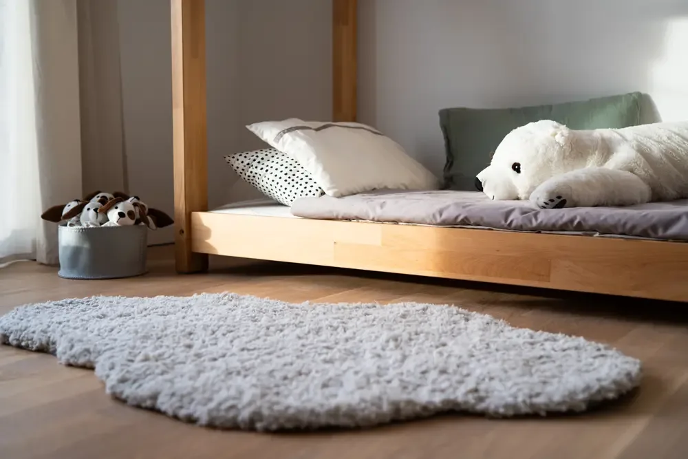 Gemütliches Kinderzimmer mit einem Holzbett, einem großen weißen Plüschtier und einem weichen, grauen Teppich. Im Hintergrund sind Kissen mit verschiedenen Mustern und eine Box mit Stoffhunden zu sehen. Diese Immobilie am Albertussee in Düsseldorf wurde von raum media professionell fotografiert.