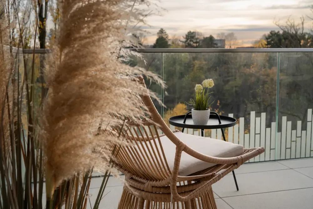 Ein gemütlicher Balkon mit einem Korbsessel, einem kleinen Tisch und einer Blume in einem Topf, umgeben von hohen Pflanzen und einer modernen Glasbrüstung, mit Blick auf den Albertussee in Düsseldorf.