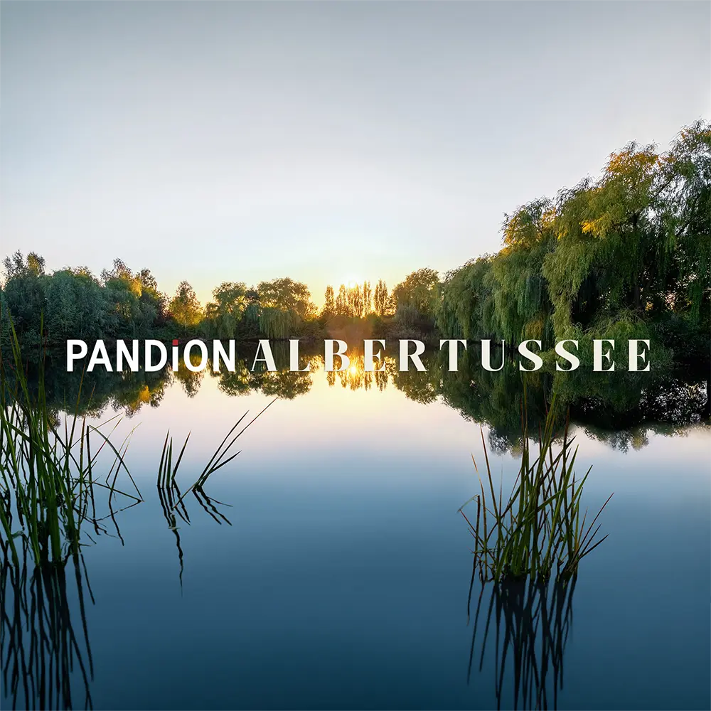 PANDION Albertussee mit ruhiger Wasseroberfläche und grünen Uferpflanzen, reflektierender Sonnenaufgang im Hintergrund.