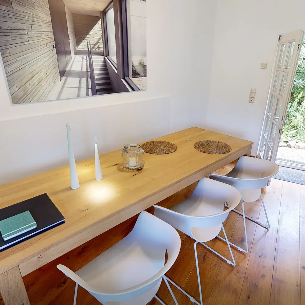 Ein heller Essbereich mit einem langen Holztisch, weißen Stühlen und dekorativen Platzdeckchen. An der Wand hängt ein großes Bild eines Treppenhauses mit viel natürlichem Licht.