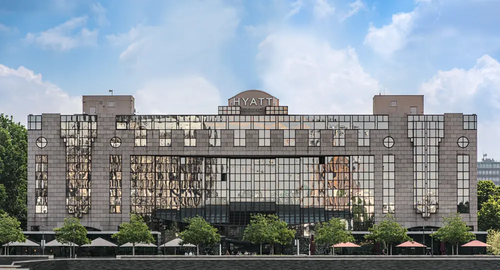 Hyatt Hotel mit moderner Architektur und großen Fensterflächen, umgeben von Bäumen und Sonnenschirmen im Vordergrund.