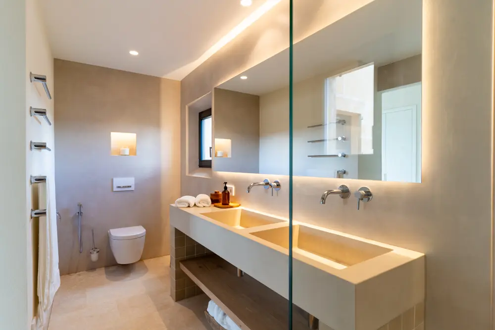 Modernes Badezimmer mit zwei Waschbecken, einem WC und einer großen Glasduschwand, beleuchtet von indirekter LED-Beleuchtung.