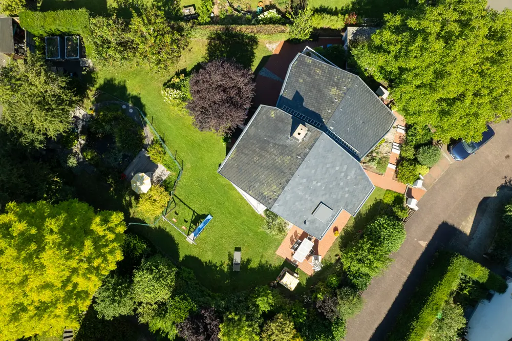 Luftaufnahme eines Hauses mit einem großen Garten, umgeben von Bäumen und Pflanzen, sowie einem Bereich mit Gartenmöbeln und einer Schaukel.