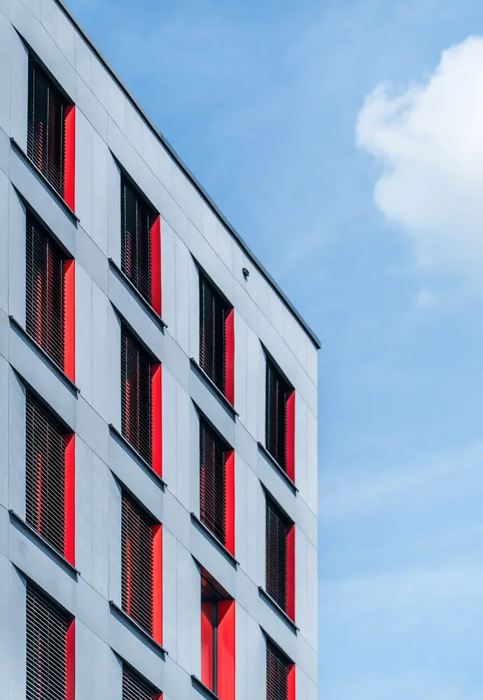 Fassade eines modernen Gebäudes mit roten Akzenten an den Fenstern vor einem blauen Himmel.