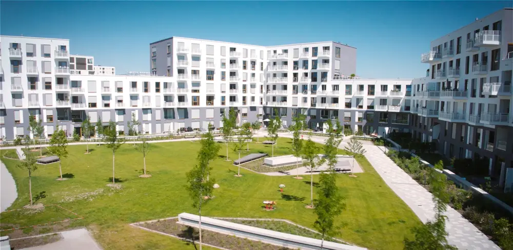 Moderne Wohnanlage mit mehreren Gebäuden, umgeben von einem gepflegten Garten mit Rasenflächen und jungen Bäumen.