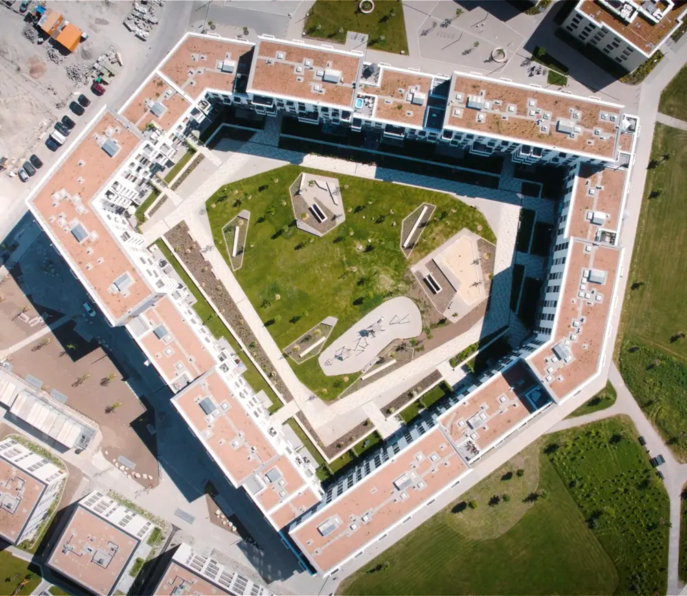 Drohnenaufnahme eines modernen Gebäudekomplexes mit einem begrünten Innenhof und verschiedenen Spielgeräten.