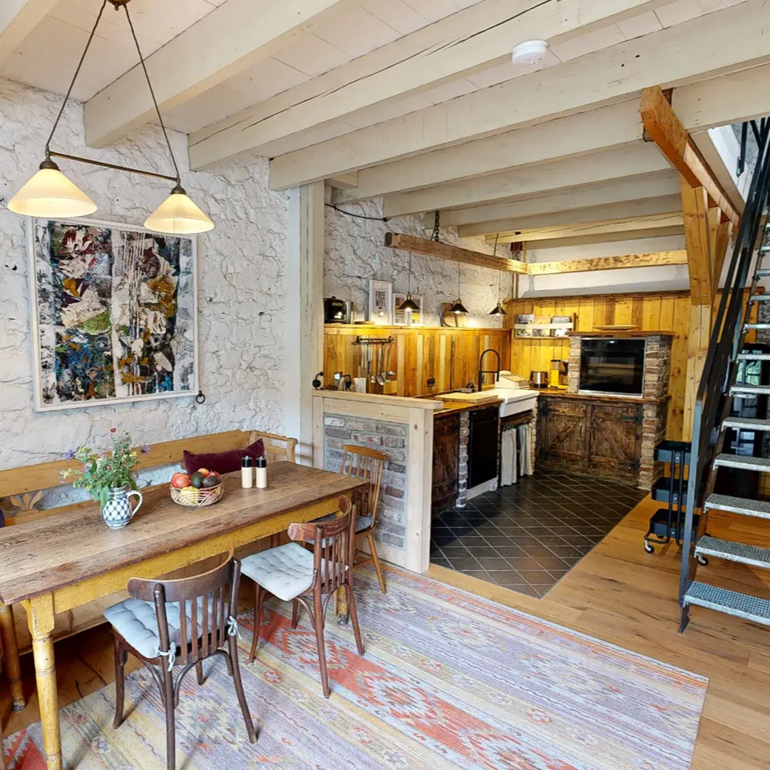 Ein heller, einladender Wohnraum mit einem Holztisch, mehreren Stühlen und einer offenen Küche im Hintergrund. Die Wände sind aus Stein, und es gibt Holzverkleidungen, die Wärme ausstrahlen. Ein Teppich verleiht dem Raum Gemütlichkeit.