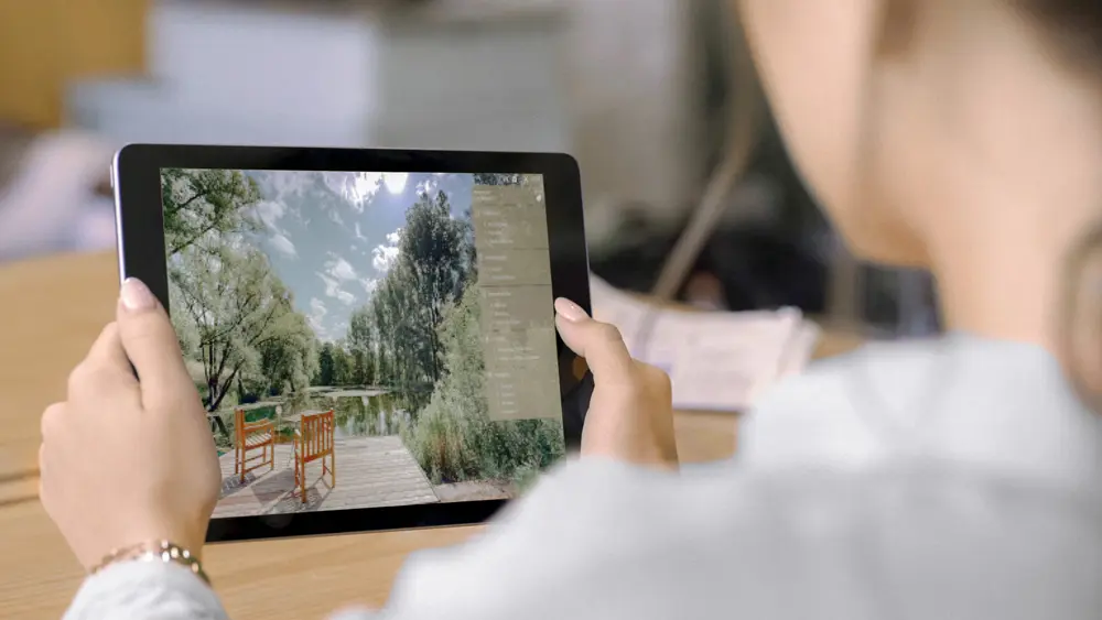 Eine Person hält ein Tablet mit beiden Händen und betrachtet eine virtuelle Tour von raum media. In der Tour sieht man eine Terrasse mit Holzboden, Tisch und Stühlen, umgeben von Bäumen und Sträuchern gerahmt von einer Menüführung